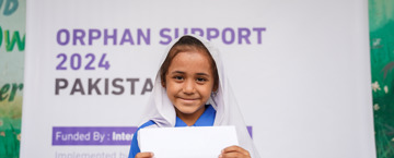 كيف اكفل يتيم في بيتي بنت تحمل ورقة أمامها لافتة كبيرة تحتوي على نص حول برنامج دعم الأيتام في باكستان لعام 2024، بتمويل من الوقف البريطاني الدولي. يشير النص إلى دور المنظمة في التمويل والتنفيذ.